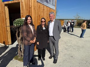 Drei Personen stehen vor einem Holzgebäude, im Hintergrund weitere Personen und eine Landschaft.