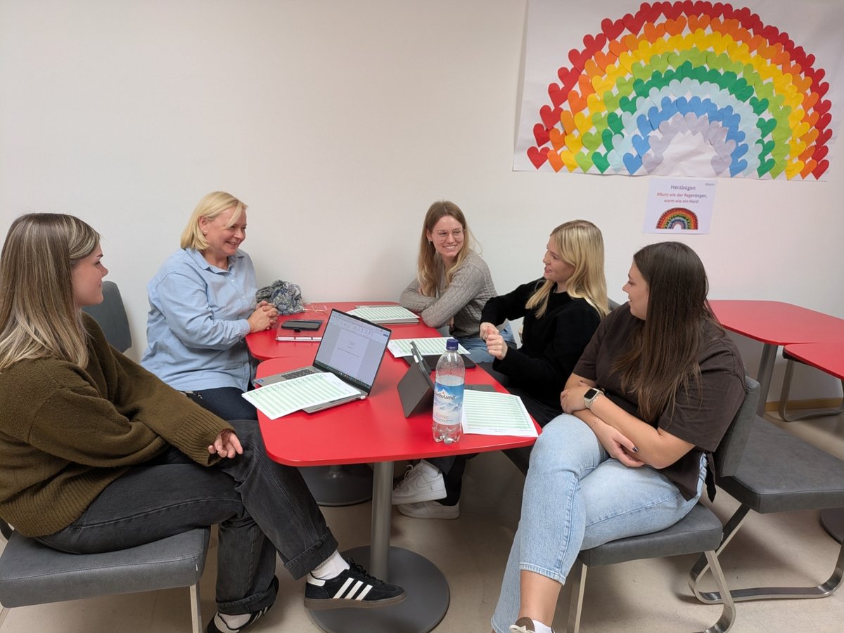 Fünf Frauen besprechen sich an einem roten Tisch in einem hellen Raum, im Hintergrund ein Regenbogen aus Herzen.