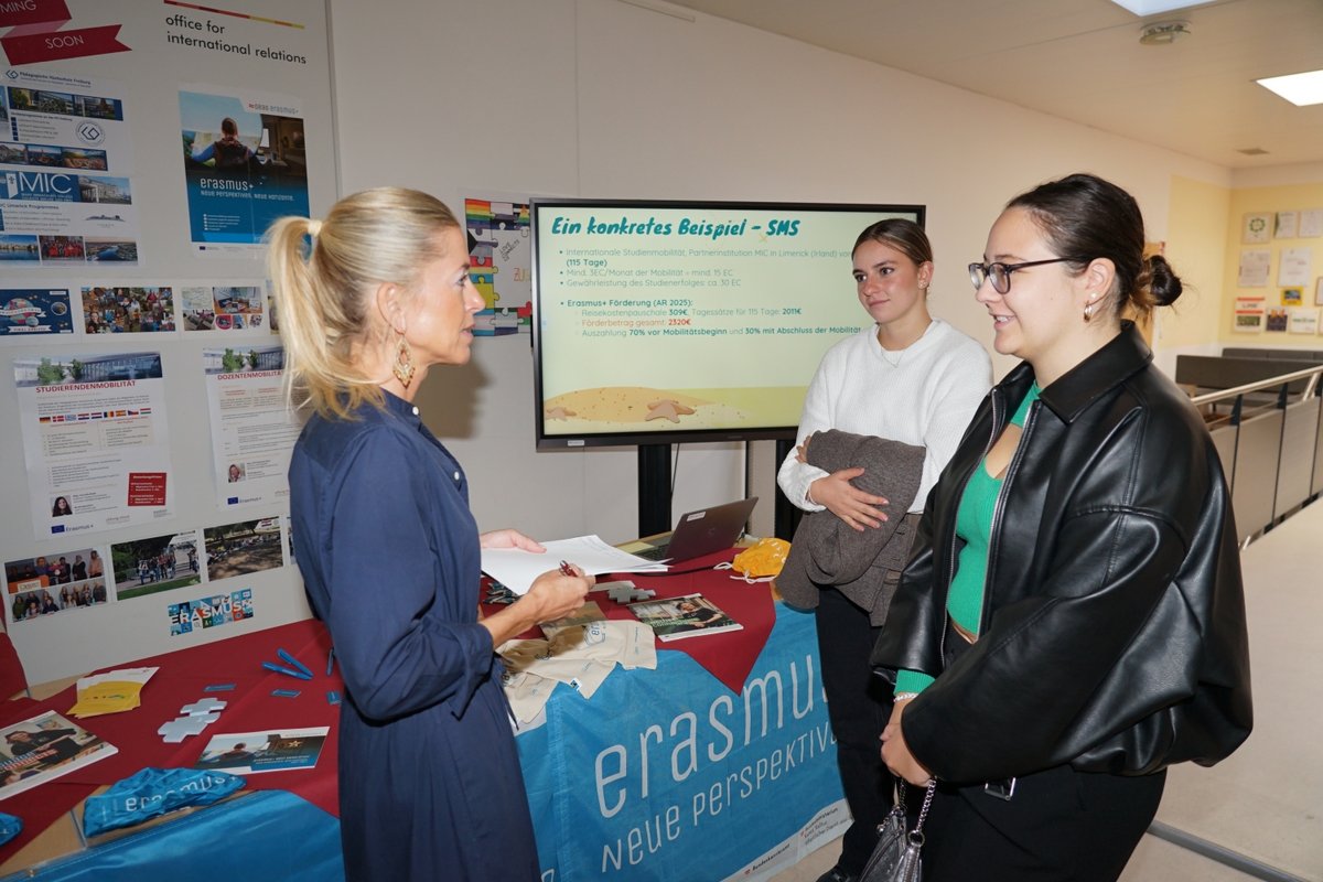Erasmus-Info: Frau im blauen Kleid erklärt zwei Studentinnen ein Poster über Auslandsaufenthalte.