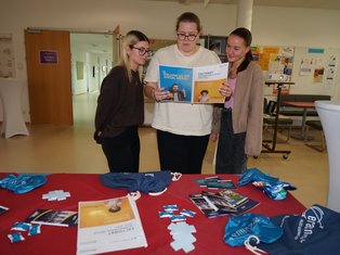 Drei junge Frauen betrachten eine Broschüre auf einem Tisch mit Informationsmaterial in einem Gebäude.